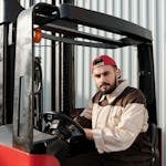 Portrait of a forklift operator inside a warehouse, wearing a uniform and cap.