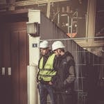 Two construction workers in safety gear standing outdoors at a building site, focused on work.